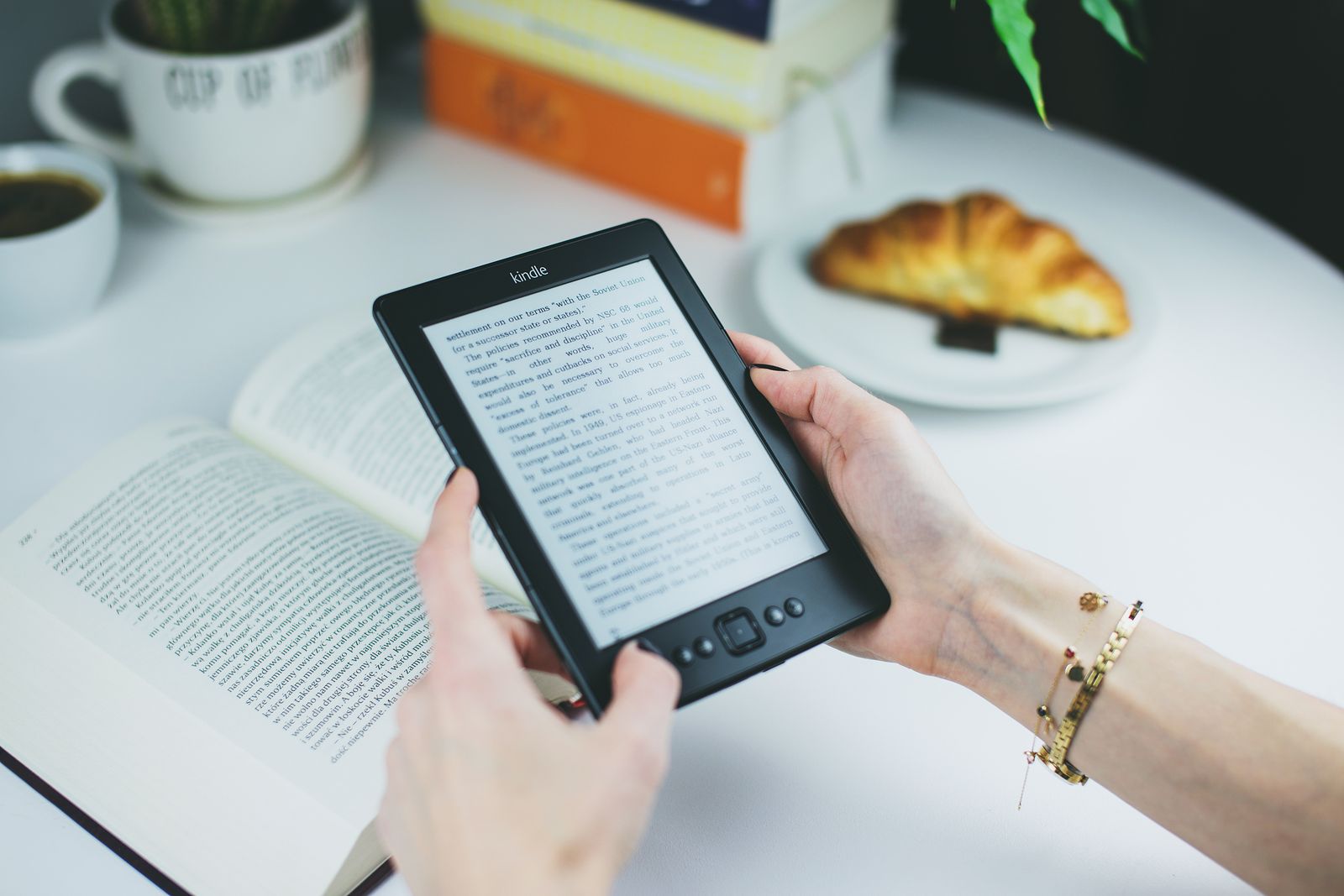 Hands holding a Kindle e-reader next to an open book and a croissant